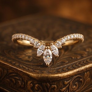 May include: A gold V-shaped ring featuring a band set with small round brilliant-cut stones and a central cluster of three marquise-cut stones. The design follows a chevron silhouette with clear, faceted stones embedded along the polished gold band.