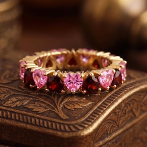 Op de afbeelding: Een gouden eternity-ring met afwisselend hartvormige roze en donkerrode edelstenen. De ring is opgebouwd uit een doorlopende rij gefacetteerde stenen in een metalen zetting, wat zorgt voor een cirkelvormig ontwerp met een herhalend patroon.