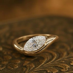 May include: A gold ring featuring a marquise-cut diamond. The ring's band has a flowing design that cradles the diamond. The ring is displayed on a decorative, antique-style box.