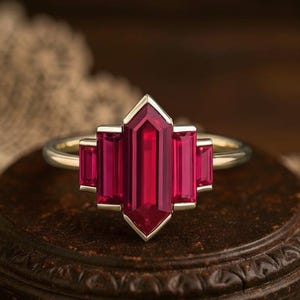 May include: A gold ring featuring a central, elongated, rectangular ruby gemstone, flanked by two smaller rectangular rubies on each side. The ring has a geometric design with a vintage aesthetic, set against a dark background.