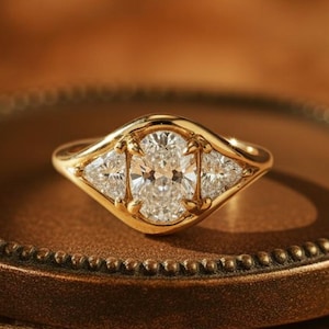 Peut inclure: Une bague en or avec une pierre centrale en diamant de taille ovale et deux pierres latérales triangulaires. La bague est sertie sur un anneau en or et est présentée sur une surface marron texturée. C'est un bijou.