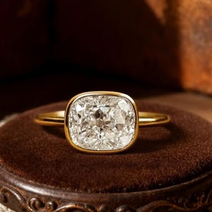Op de afbeelding: Een gouden ring met een grote, vierkant geslepen diamant. De diamant is gezet in een bezel setting en zit op een gouden band. De ring wordt getoond op een bruin fluwelen oppervlak met een decoratief kanten kleedje.
