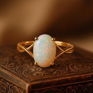 May include: A gold ring featuring a central oval-shaped opal gemstone with iridescent flashes of colour. The band is crafted from polished gold and splits into a delicate, crossed design on both sides of the prong-set opal stone.