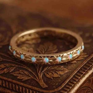 May include: A gold-toned band ring featuring a repeating starburst pattern engraved along the outer surface. Each starburst center is set with a small, round, iridescent opal gemstone, creating a continuous circular design around the entire ring.