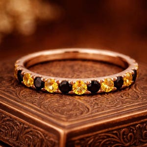 May include: A rose gold band ring featuring an alternating pattern of round-cut yellow and black gemstones set in a shared prong setting. The ring is circular and displays a continuous row of stones around the entire circumference of the metal band.