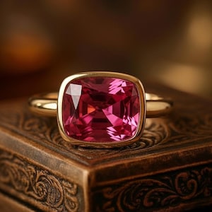 Pode incluir: Um anel de ouro com uma grande pedra rosa com corte almofada. O anel tem uma moldura simples e uma banda de ouro lisa e polida. O anel é exibido em uma caixa de madeira escura e ornamentada.