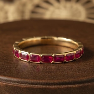 May include: A gold band ring featuring a continuous row of rectangular, bezel-set red rubies. The ring has a polished metallic finish and a uniform design, showcasing a series of vibrant, deep red gemstones set securely within the gold metal frame.