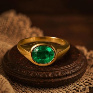 Puede incluir: Un anillo de oro con una gran esmeralda de talla ovalada. El anillo tiene un diseño clásico y la esmeralda es de color verde intenso. El anillo se exhibe sobre una superficie marrón oscuro y texturizada.