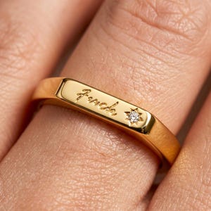 May include: A gold-toned ring featuring a flat rectangular top surface engraved with the word "fuck" in a cursive script, accompanied by a small, clear, round-cut gemstone set within a star-shaped indentation on the right side of the band.