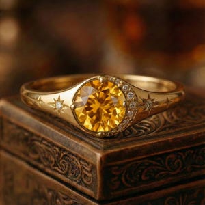 May include: A gold ring featuring a large, round, yellow gemstone. The ring has a celestial design with stars and small diamonds. The ring is set on a dark, ornate wooden box, creating a warm, inviting aesthetic.