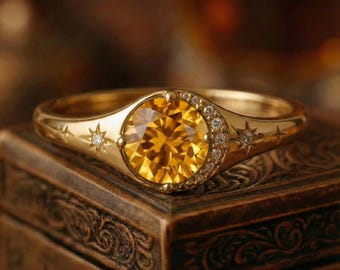 May include: A gold ring featuring a large, round, yellow gemstone. The ring has a celestial design with stars and small diamonds. The ring is set on a dark, ornate wooden box, creating a warm, inviting aesthetic.