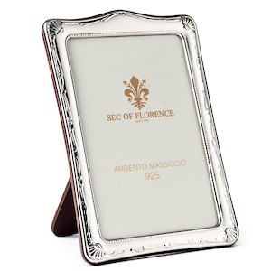 May include: A silver picture frame with a decorative edge. The frame has a white interior with text that reads "SEC OF FLORENCE" and "ARGENTO MASSICCIO 925". It is supported by a brown stand.