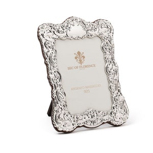 May include: A silver picture frame with an ornate design. The frame has a stand on the back and says "SEC OF FLORENCE ARGENTIO MASSICCIO 925" on the front.