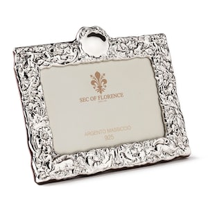 May include: A silver picture frame with an ornate design. The frame has a raised, rounded top and a small inscription that reads "SEC OF FLORENCE since 1984" and "ARGENTO MASSICCIO 925".