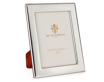 May include: Silver picture frame with a textured design and a wooden stand. The frame has the text "SEC OF FLORENCE" and "ARGENTO MASSICCIO 925" printed on it.