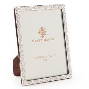 May include: A silver picture frame with a decorative border of small circles. It has a wooden backing and stand. The text "SEC OF FLORENCE" and "ARGENTO MASSICCIO 925" are visible on the white background. The frame is rectangular.