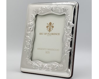 Puede incluir: Un marco de fotos plateado con un diseño floral de rosas alrededor del borde. El marco tiene una parte trasera marrón oscuro y presenta el texto "SEC OF FLORENCE" y "ARGENTO MASSICCIO 925".