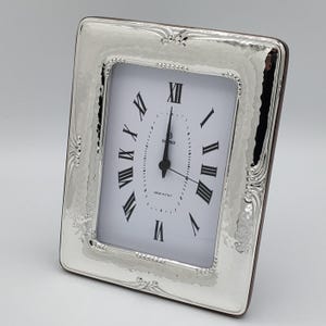 May include: A rectangular silver-toned clock with a white face and black Roman numerals. The clock has a hammered texture on the frame and decorative flourishes on the corners. The clock reads 12:00. The words "MADE IN ITALY" are printed on the clock face.