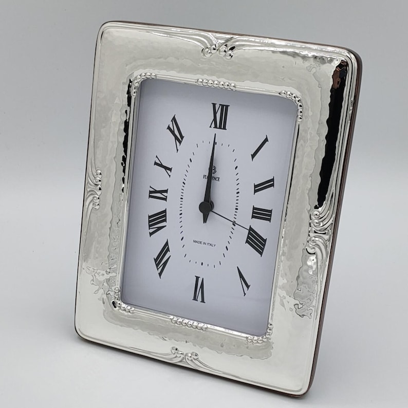 May include: A rectangular silver-toned clock with a white face and black Roman numerals. The clock has a hammered texture on the frame and decorative flourishes on the corners. The clock reads 12:00. The words "MADE IN ITALY" are printed on the clock face.