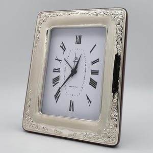 May include: Silver-toned rectangular clock with a white face and black Roman numerals. The clock has a decorative, ornate frame and black clock hands. The clock face reads "Made in Italy".