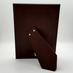 May include: A dark brown wooden picture frame with a stand on the back. The frame has a simple design and is made of high-quality wood.