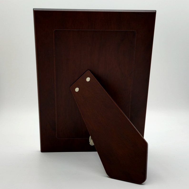 May include: A dark brown wooden picture frame with a stand on the back. The frame has a simple design and is made of high-quality wood.