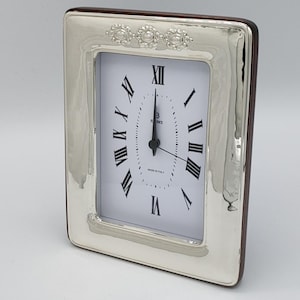 May include: A rectangular silver-toned clock with a white face and black Roman numerals. The clock has a decorative floral design at the top and a dark brown wooden backing. The clock reads 12:00. The words "MADE IN ITALY" are printed on the clock face.