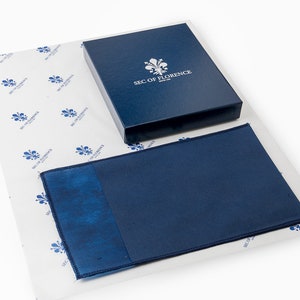 Pode incluir: Uma carteira de couro azul marinho com acabamento texturizado, apresentada numa caixa a condizer. A caixa &eacute; estampada com um emblema floral e o texto "SEC OF FLORENCE". A carteira repousa sobre papel de seda estampado.
