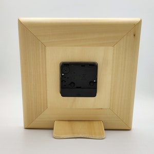 May include: The back of a wooden clock with a black clock mechanism. The clock has a stand on the bottom.
