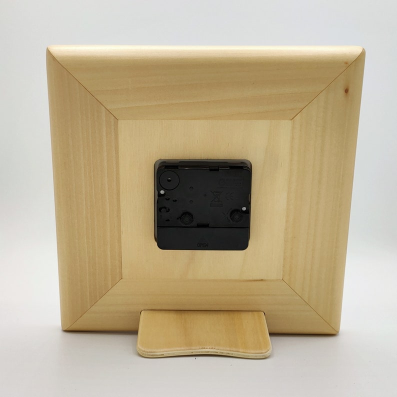 May include: The back of a wooden clock with a black clock mechanism. The clock has a stand on the bottom.