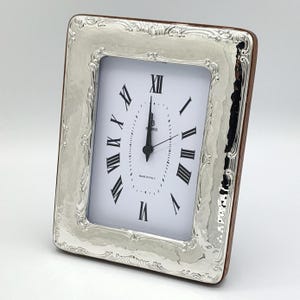 May include: Silver-toned rectangular clock with a white face and black Roman numerals. The clock has a decorative, hammered-look silver frame and a brown wooden backing. The clock reads 12:00. The words "MADE IN ITALY" are printed on the clock face.
