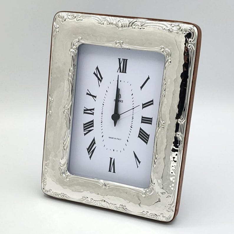 May include: Silver-toned rectangular clock with a white face and black Roman numerals. The clock has a decorative, hammered-look silver frame and a brown wooden backing. The clock reads 12:00. The words "MADE IN ITALY" are printed on the clock face.