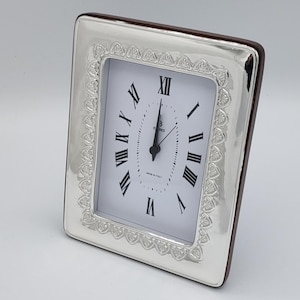 May include: A rectangular silver-toned clock with a white face and black Roman numerals. The clock has a decorative border and a dark brown wooden backing. The clock face reads "Florence" and "Made in Italy".