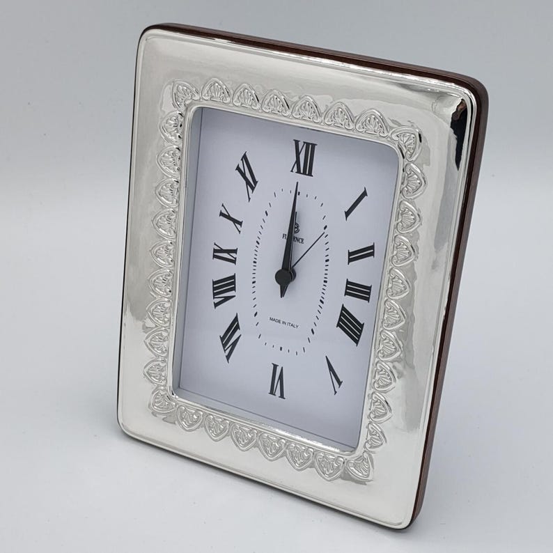 May include: A rectangular silver-toned clock with a white face and black Roman numerals. The clock has a decorative border and a dark brown wooden backing. The clock face reads "Florence" and "Made in Italy".