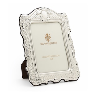 May include: A silver picture frame with an ornate design. The frame has a stand on the back and says "SEC of FLORENCE since 1980" and "ARGENTO MASSICCIO 925" on the front.