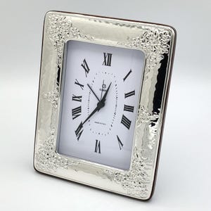 K&ouml;nnte beinhalten: Eine rechteckige Uhr in Silberoptik mit wei&szlig;em Zifferblatt und schwarzen r&ouml;mischen Ziffern. Die Uhr hat einen dekorativen floralen Rahmen und eine braune R&uuml;ckseite. Auf dem Zifferblatt steht "Made in Italy".