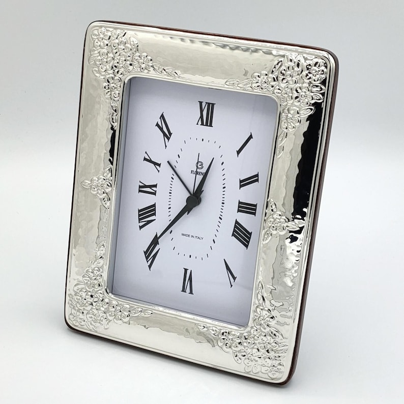 K&ouml;nnte beinhalten: Eine rechteckige Uhr in Silberoptik mit wei&szlig;em Zifferblatt und schwarzen r&ouml;mischen Ziffern. Die Uhr hat einen dekorativen floralen Rahmen und eine braune R&uuml;ckseite. Auf dem Zifferblatt steht "Made in Italy".