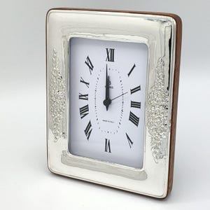 Pu&ograve; includere: Orologio rettangolare argentato con quadrante bianco e numeri romani neri. L'orologio ha un retro in legno e decorazioni floreali sui lati. Le lancette indicano le 12:00.