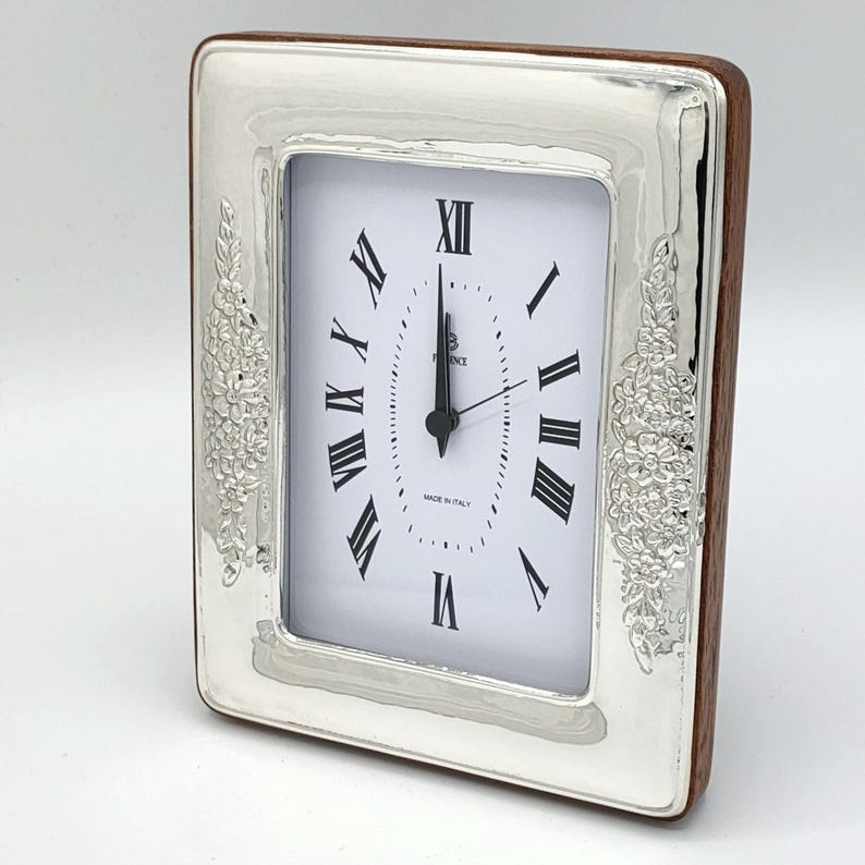Pu&ograve; includere: Orologio rettangolare argentato con quadrante bianco e numeri romani neri. L'orologio ha un retro in legno e decorazioni floreali sui lati. Le lancette indicano le 12:00.