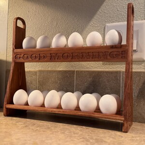 May include: A wooden egg rack with two tiers, holding 14 white eggs. The rack is made of dark wood and has the words "GOOD MORNING!" carved into the front. The rack has a handle on each side. The rack is on a countertop.