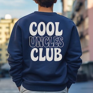 Könnte beinhalten: Ein marineblaues Sweatshirt mit dem weißen Aufdruck "COOL UNCLES CLUB" mit weißer Umrandung. Die Person, die das Sweatshirt trägt, hat braunes Haar und steht vor einem verschwommenen urbanen Hintergrund.