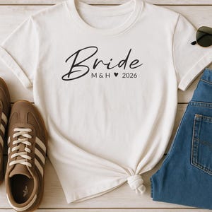 Könnte beinhalten: Ein weißes T-Shirt mit dem Wort "Bride" in Schreibschrift und "M & H ♥ 2026" in Schwarz gedruckt. Das Shirt wird mit braunen Turnschuhen, einer blauen Jeans und einer Sonnenbrille präsentiert.
