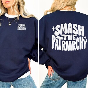 Könnte beinhalten: Marineblaues Sweatshirt mit dem weißen Text "SMASH THE PATRIARCHY" auf der Vorder- und Rückseite. Die Vorderseite hat eine kleinere Version des Textes. Das Sweatshirt hat einen weißen Kragen.