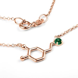 Rose Gold necklace in polished finish, featuring the molecule of the Dopamine hormone. The atom on the right end of the molecule has an emerald set in it. The molecule stands between a thin link chain also in rose gold.