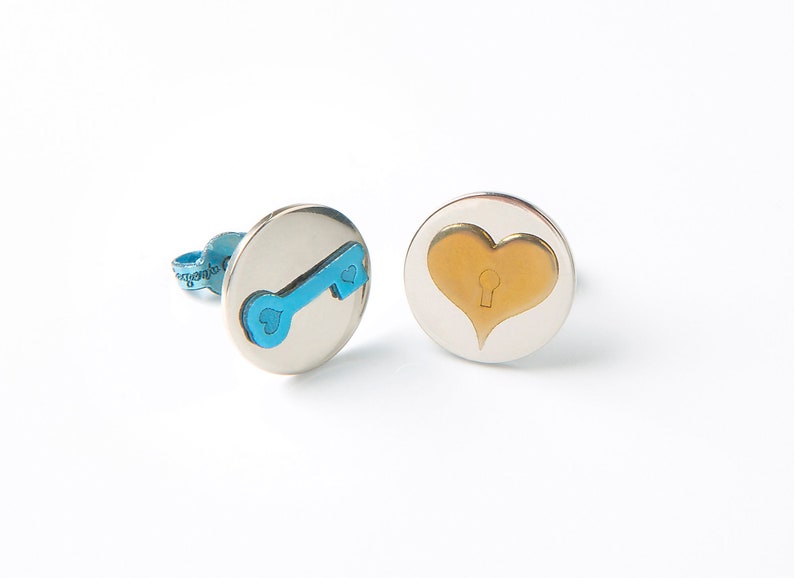 Round stud earrings made with Titanium. Their base is silver colored, on one of the studs there is a key in Sky Blue color and on the other a heart in Bronze color.