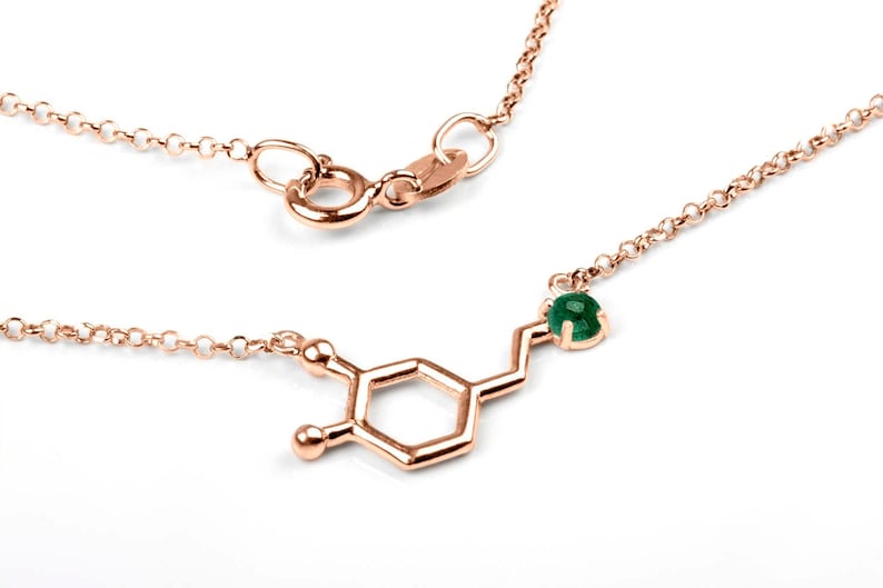 Rose Gold necklace in polished finish, featuring the molecule of the Dopamine hormone. The atom on the right end of the molecule has an emerald set in it. The molecule stands between a thin link chain also in rose gold.