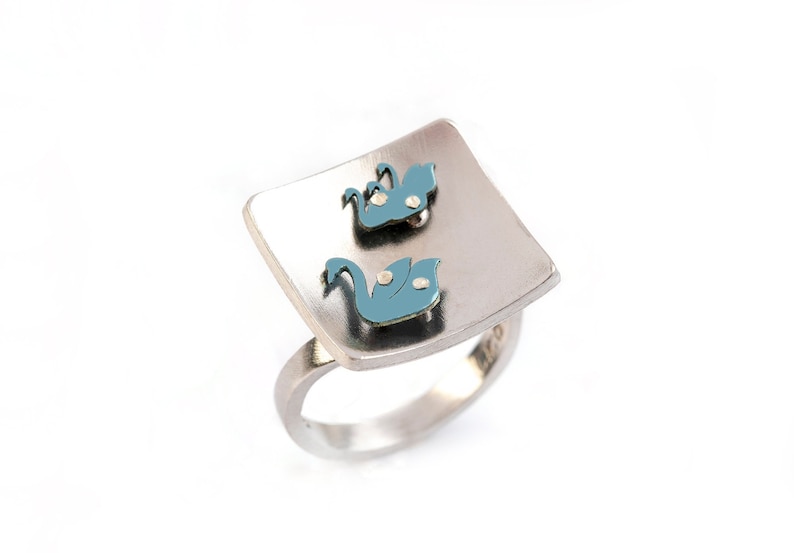 Square silver ring with a colored theme of titanium parts on its top. It has a bright mat finish and flat band. The theme is a Swan floating next to three of its baby swans, all in silver blue color. Two tiny silver flat pins hold each titanium part.