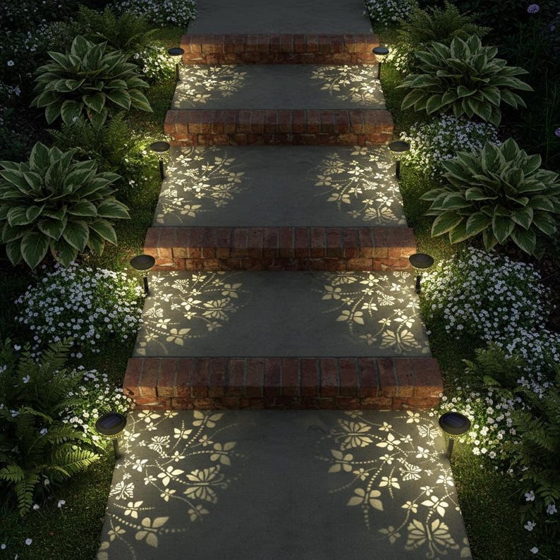 May include: Outdoor pathway with brick steps and solar lights. The steps are illuminated with a floral pattern. Lush green plants and white flowers border the pathway, creating a serene garden scene. The lights cast a warm glow.