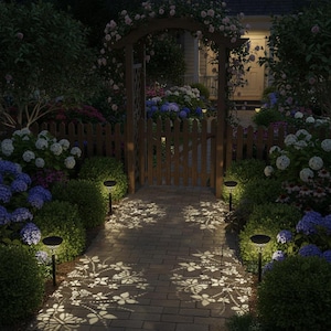 May include: A nighttime garden scene with a brick pathway illuminated by solar lights. The lights cast a floral pattern on the path. A wooden gate and archway are visible, surrounded by lush greenery and colorful flowers.