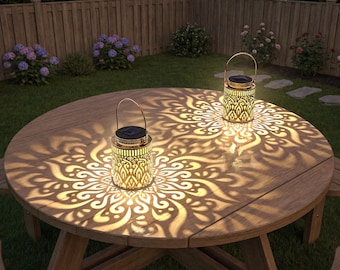 May include: Two cylindrical metal lanterns with intricate cut-out patterns sit on a round wooden table. Each lantern features a top-mounted solar panel and a wire handle, casting a bright, symmetrical floral light projection onto the table surface.